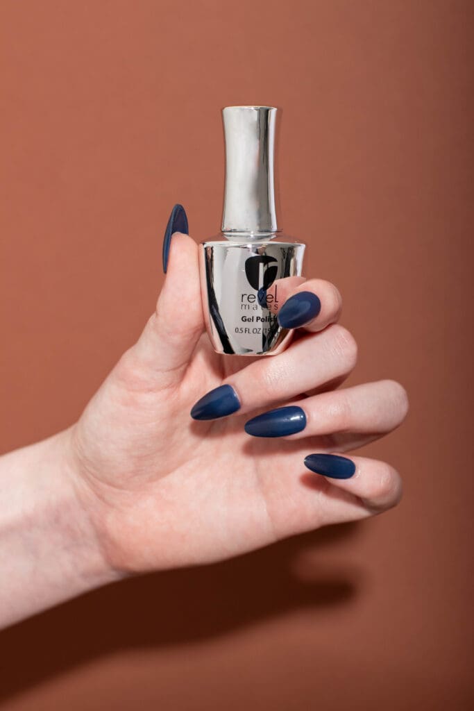 A female model holding a gel polish bottle with navy blue nails with a brown background.